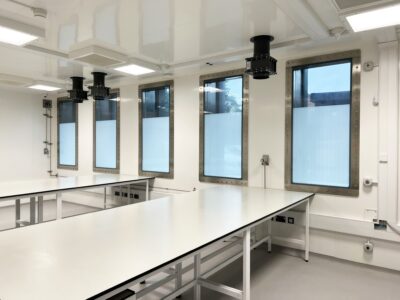 A bright, modern laboratory equipped with a High Containment APR Vision Panel, white walls, and large windows letting in natural light. Long, empty tables and ceiling vents contribute to a clean, sterile atmosphere.