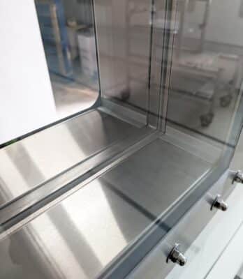 Close-up of a High Containment APR Vision Panel integrated into a metallic corner structure with glass panels reflecting light. The blurred industrial background complements the modern, sleek design.