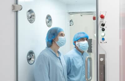 Two cleanroom personnel in gowns and face masks standing inside a PBSC air shower with high velocity nozzles and interlocked door controls prior to cleanroom entry