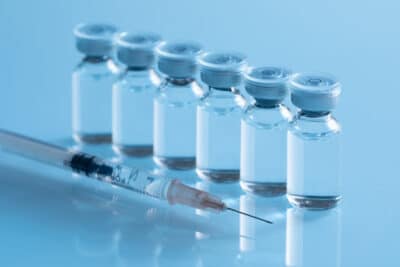 Row of pharmaceutical vaccine vials with blue caps alongside a syringe, representing drug manufacturing environments where PBSC cleanroom solutions support contamination control