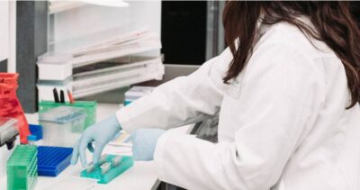 Capricor Therapeutics laboratory researcher in white lab coat and blue gloves handling sample vials and microplate trays at a controlled environment workstation, representing the precision workflows PBSC decontamination systems are designed to support
