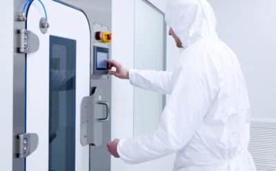 Laboratory operative in full white cleanroom suit operating a touchscreen control panel on a high containment pass-through hatch, demonstrating PBSC equipment designed for safe, intuitive use
