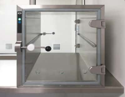 PBSC wall-mounted transfer hatch with open toughened glass door showing passive mechanical interlock bar, stainless steel frame, green status indicator and glass shelf installed in a cleanroom