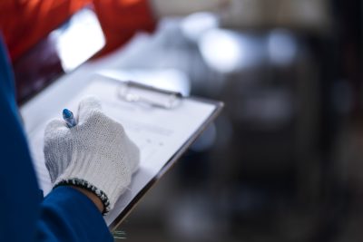 Close-up of a gloved engineer’s hand holding a pen and clipboard during a quality control check inside a controlled environment or cleanroom facility.