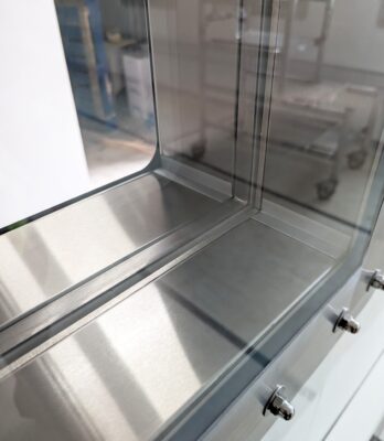 Close-up of a High Containment APR Vision Panel integrated into a metallic corner structure with glass panels reflecting light. The blurred industrial background complements the modern, sleek design.