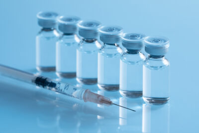 Row of pharmaceutical vaccine vials with blue caps alongside a syringe, representing drug manufacturing environments where PBSC cleanroom solutions support contamination control