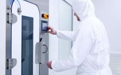 Laboratory operative in full white cleanroom suit operating a touchscreen control panel on a high containment pass-through hatch, demonstrating PBSC equipment designed for safe, intuitive use
