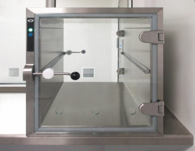 PBSC wall-mounted transfer hatch with open toughened glass door showing passive mechanical interlock bar, stainless steel frame, green status indicator and glass shelf installed in a cleanroom